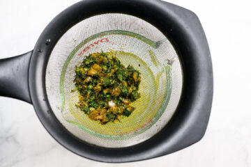 Metal strainer containing wet, chopped green and yellow plant material, placed over a black container on a white surface.