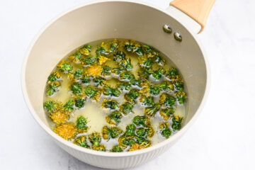 A beige pan filled with water and fresh dandelion flowers, shown on a white surface.
