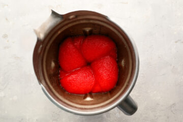 Whole peeled tomatoes in their juice are placed inside a blender jar, viewed from above on a light, textured surface.