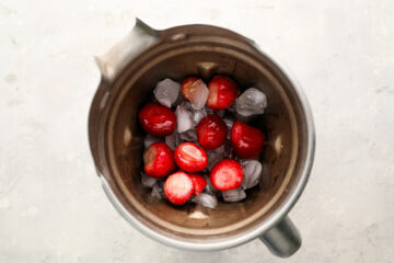 Whole strawberries and ice cubes inside a metal blender jar, viewed from above on a light surface.