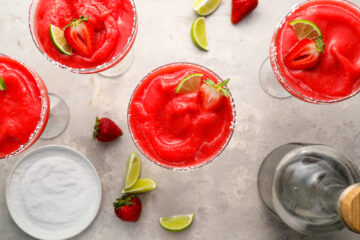Four glasses of frozen strawberry margaritas garnished with lime and strawberry slices, surrounded by lime wedges, strawberries, salt, and a bottle on a light surface.