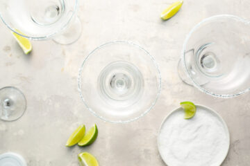 Three empty margarita glasses with salted rims, lime wedges on the table, and a plate of salt seen from above on a light surface.