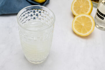 A glass of lemonade on a marble surface with lemon halves and a bottle in the background.