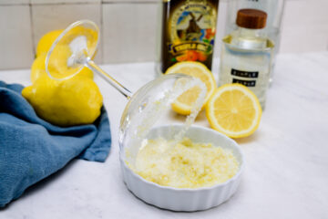 A cocktail glass rimmed with lemon sugar rests on a dish. Lemons, a blue cloth, olive oil, and a bottle of clear liquid are in the background on a marble surface.