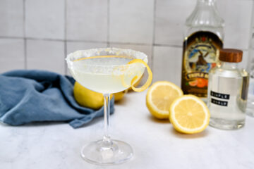 A cocktail in a coupe glass with a sugared rim and lemon twist garnish, surrounded by lemons, a blue cloth, and two liquor bottles on a white countertop.
