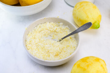 A bowl filled with a coarse lemon sugar mixture sits on a white surface, with a spoon in the bowl and whole lemons nearby.