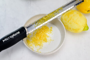 A Microplane zester rests on a bowl containing lemon zest, with whole lemons nearby on a white surface.