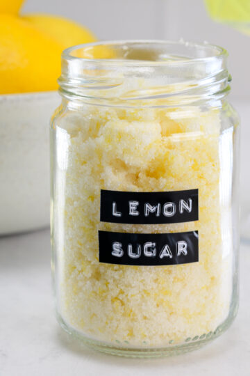 A glass jar labeled “lemon sugar” is filled with yellow sugar, with a bowl of lemons and a glass of green liquid in the background.