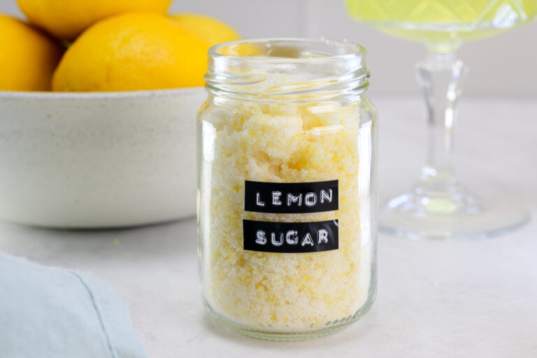A glass jar labeled “lemon sugar” is filled with yellow sugar, with a bowl of lemons and a glass of green liquid in the background.