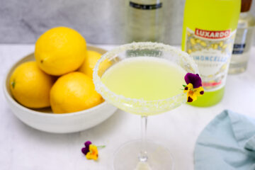 A cocktail glass with a sugar rim filled with a yellow drink, garnished with an edible flower, next to a bowl of lemons and a bottle of Luxardo Limoncello.