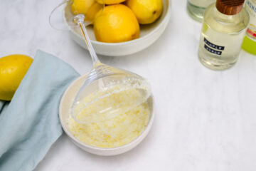 A cocktail glass is rimmed with a lemon-sugar mixture in a bowl, next to whole lemons, a folded blue cloth, and bottles of ingredients on a white surface.