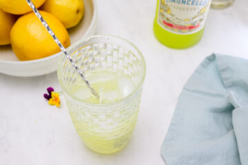 A glass of limoncello cocktail with ice and a metal straw sits on a white surface near a bottle of limoncello, a bowl of lemons, a blue napkin, and a small flower.