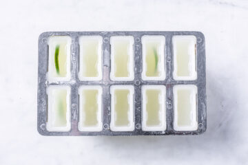 A metal popsicle mold filled with a pale yellow liquid, some sections containing small green pieces, viewed from above on a white surface.
