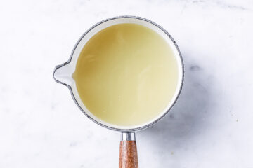 A saucepan with a light yellow liquid sits on a white marble surface, viewed from above.