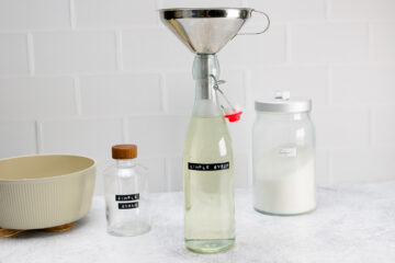A glass bottle labeled Simple Syrup being filled with liquid through a metal funnel, with a jar of sugar, a bowl, and a small bottle in the background.