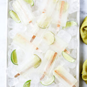 Rectangular tray filled with ice cubes, lime wedges, and several clear popsicles with wooden sticks, arranged on a white surface.