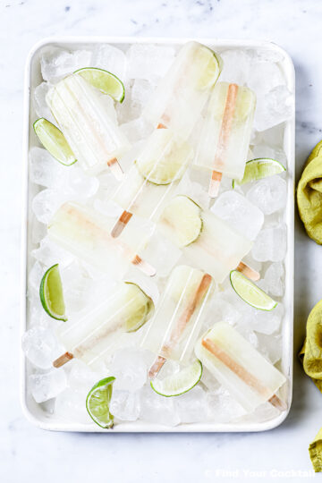Rectangular tray filled with ice cubes, lime wedges, and several clear popsicles with wooden sticks, arranged on a white surface.