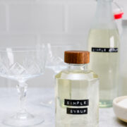 A glass bottle labeled Simple Syrup with a wooden cap sits next to a glass, a larger bottle, and a towel on a light countertop.