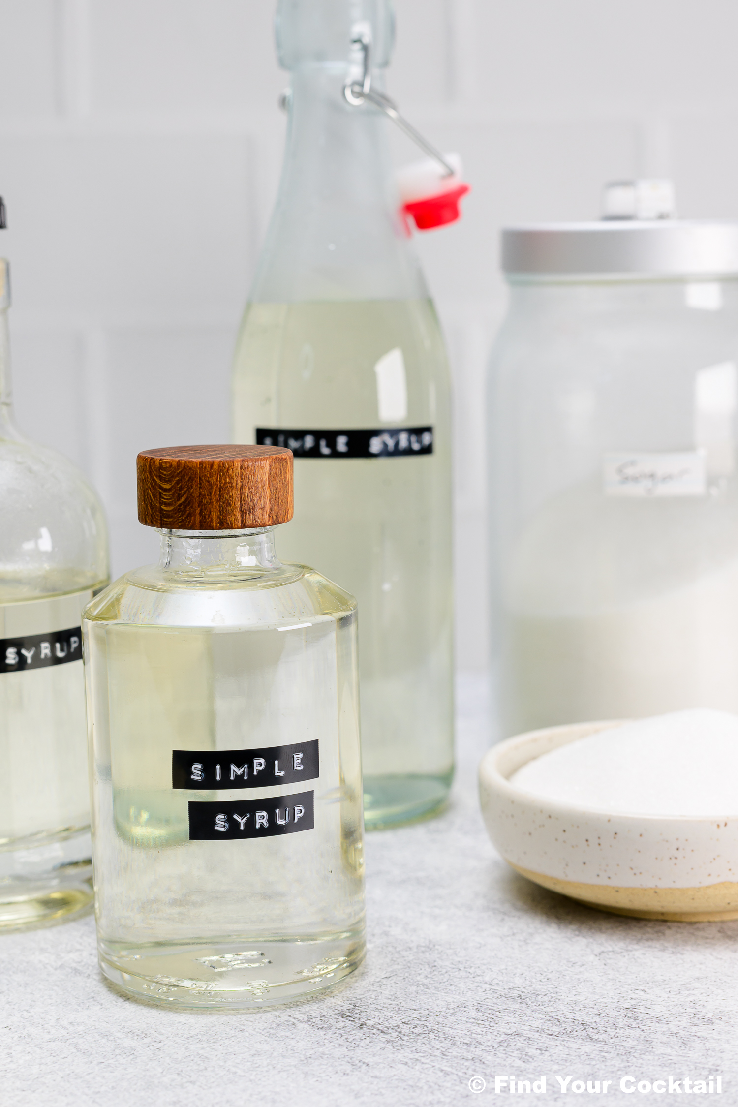 Clear glass bottles labeled Simple Syrup and a bowl of sugar are arranged on a white surface with a jar of sugar in the background.