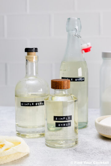 Three glass bottles labeled Simple Syrup are filled with clear liquid and placed on a light countertop with a bowl of sugar and a yellow cloth nearby.