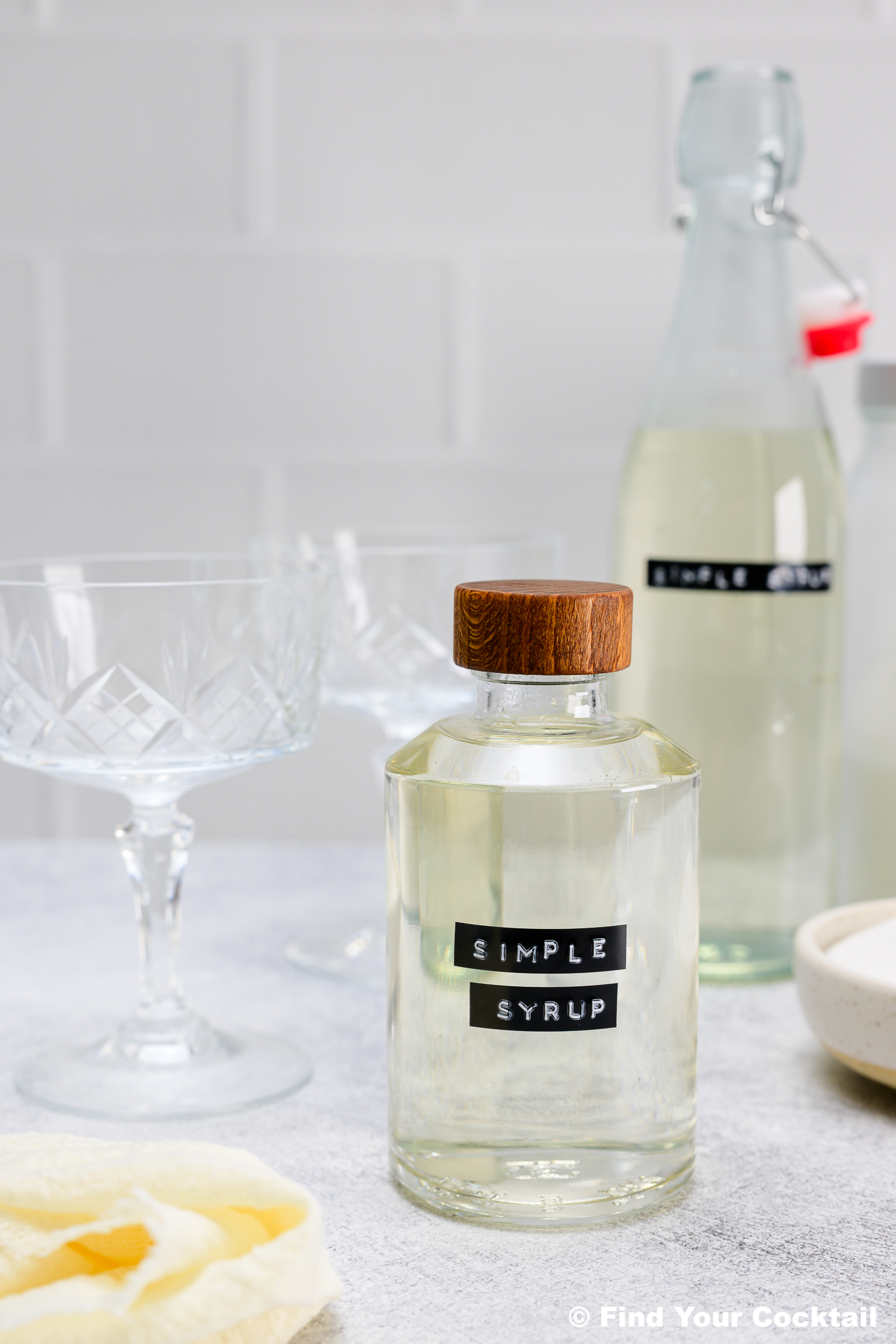 A glass bottle labeled Simple Syrup with a wooden cap sits next to a glass, a larger bottle, and a towel on a light countertop.