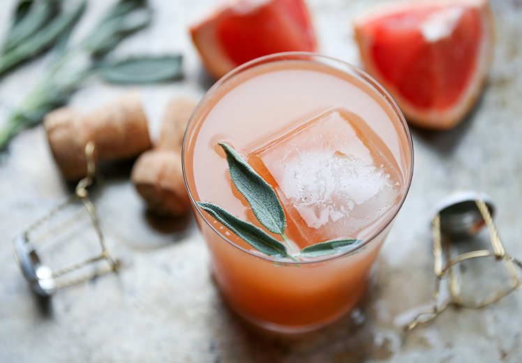 A glass of pink grapefruit cocktail with ice and a sage leaf garnish, surrounded by corks, metal cages, and grapefruit wedges.