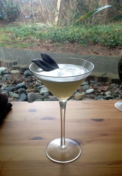 A martini glass with a light cocktail garnished with two sage leaves sits on a wooden table, with a garden visible in the background.