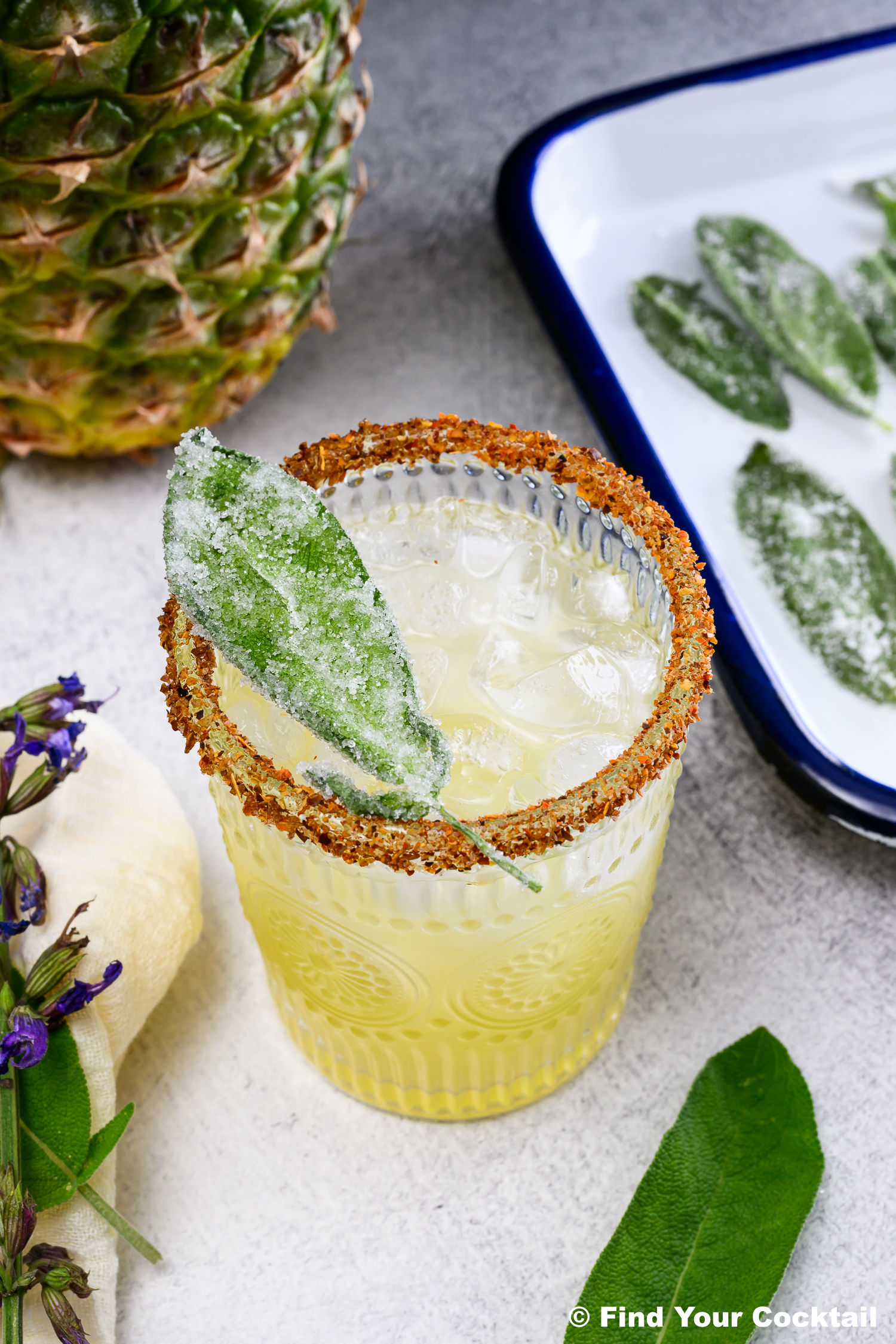 A yellow cocktail with ice in a glass rimmed with spice, garnished with a sugared sage leaf. Pineapple and sugared sage leaves are visible in the background.