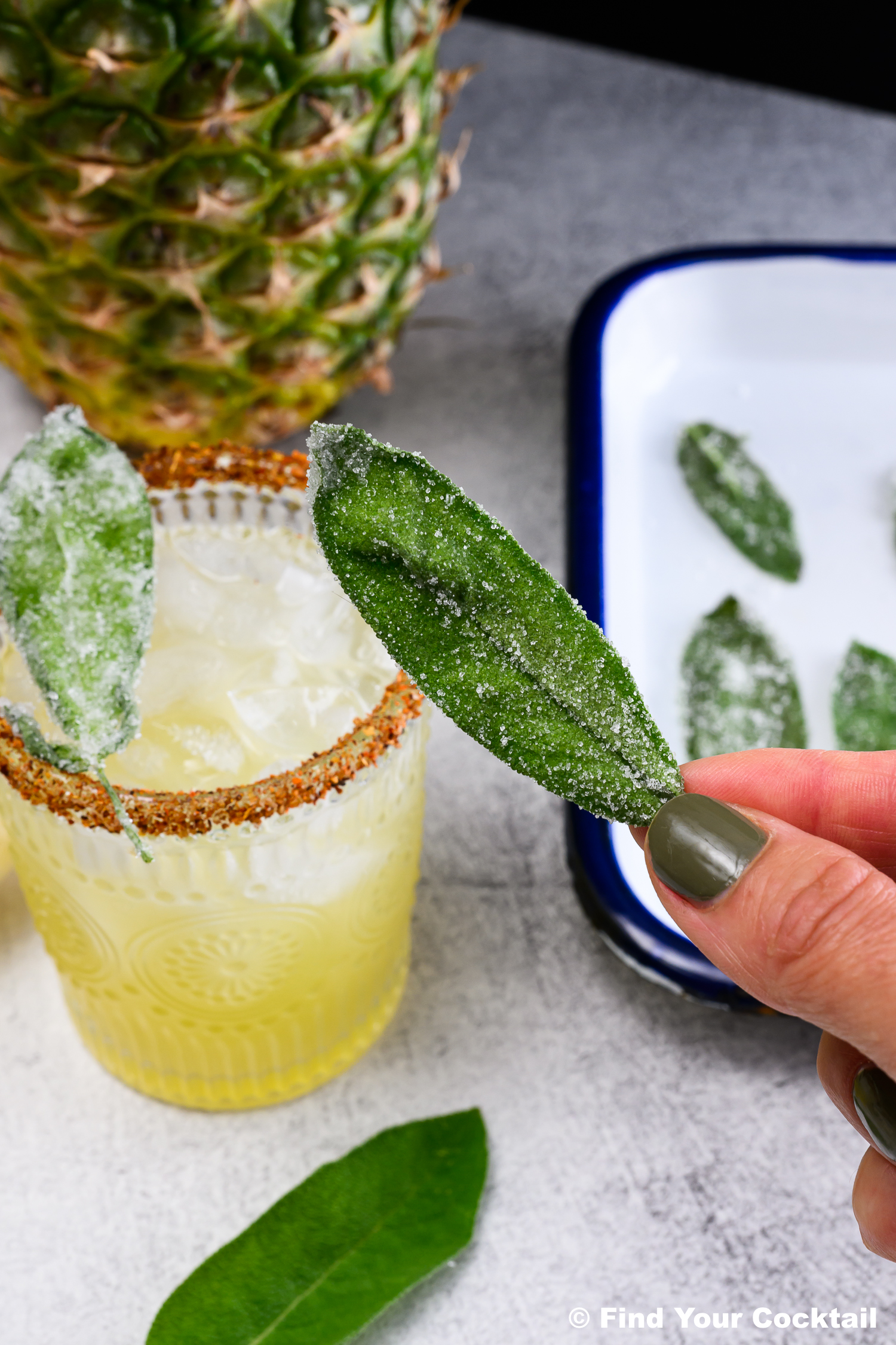 A hand holds a sugared sage leaf above a cocktail with a spiced rim; more sugared leaves and a pineapple are in the background.