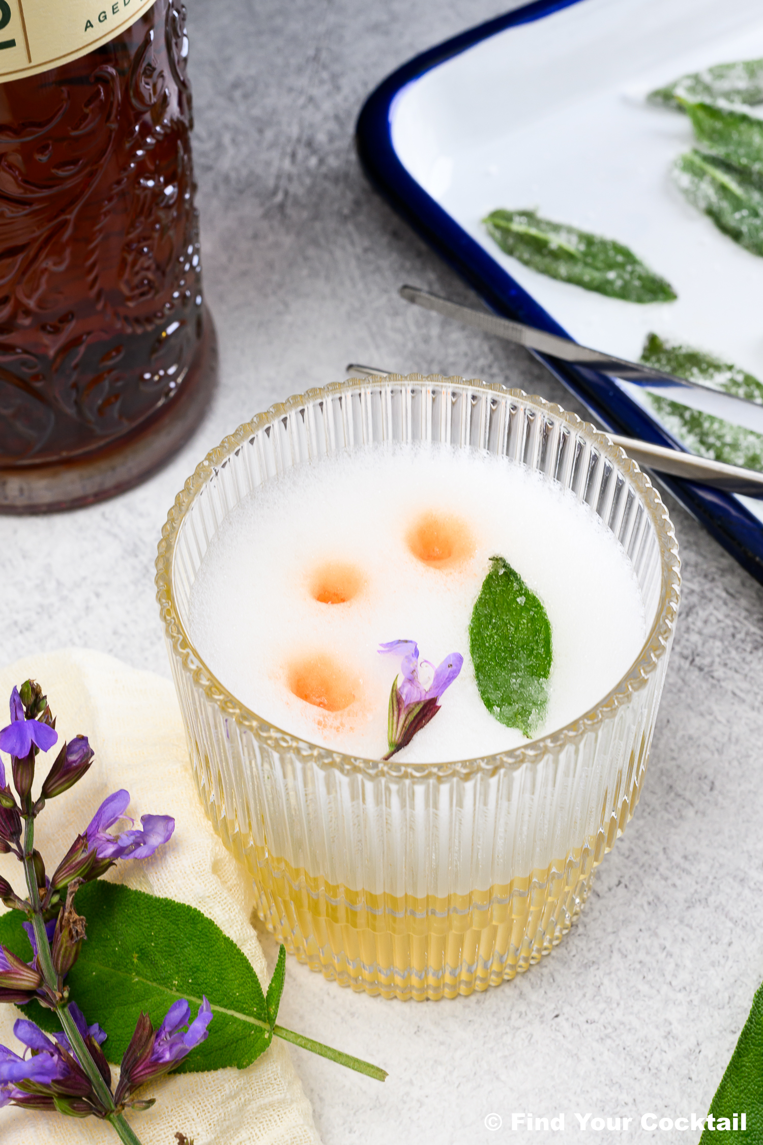 A glass of frothy cocktail garnished with a sage leaf, three drops of bitters, and a purple flower, with a tray of sage leaves and a bottle in the background.