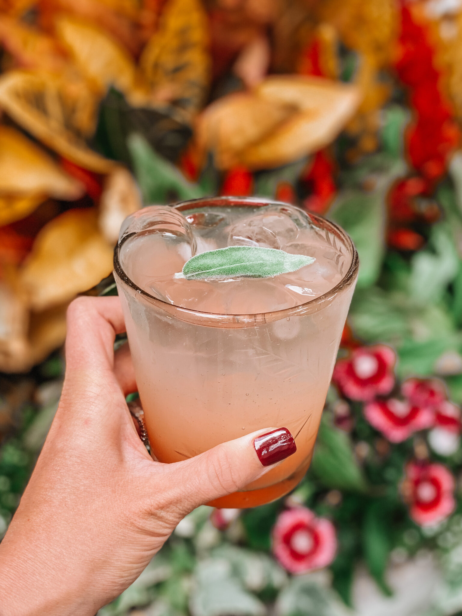 A hand with red nail polish holds a glass of iced pink beverage garnished with a green leaf, with blurred flowers and foliage in the background.