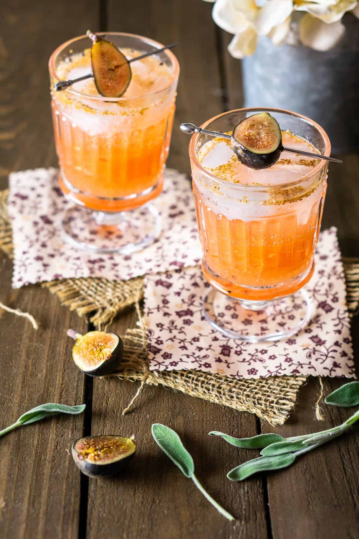 Two glasses of orange-colored cocktails topped with halved figs and brown sugar on spoons, set on patterned napkins with fresh figs and sage leaves nearby.