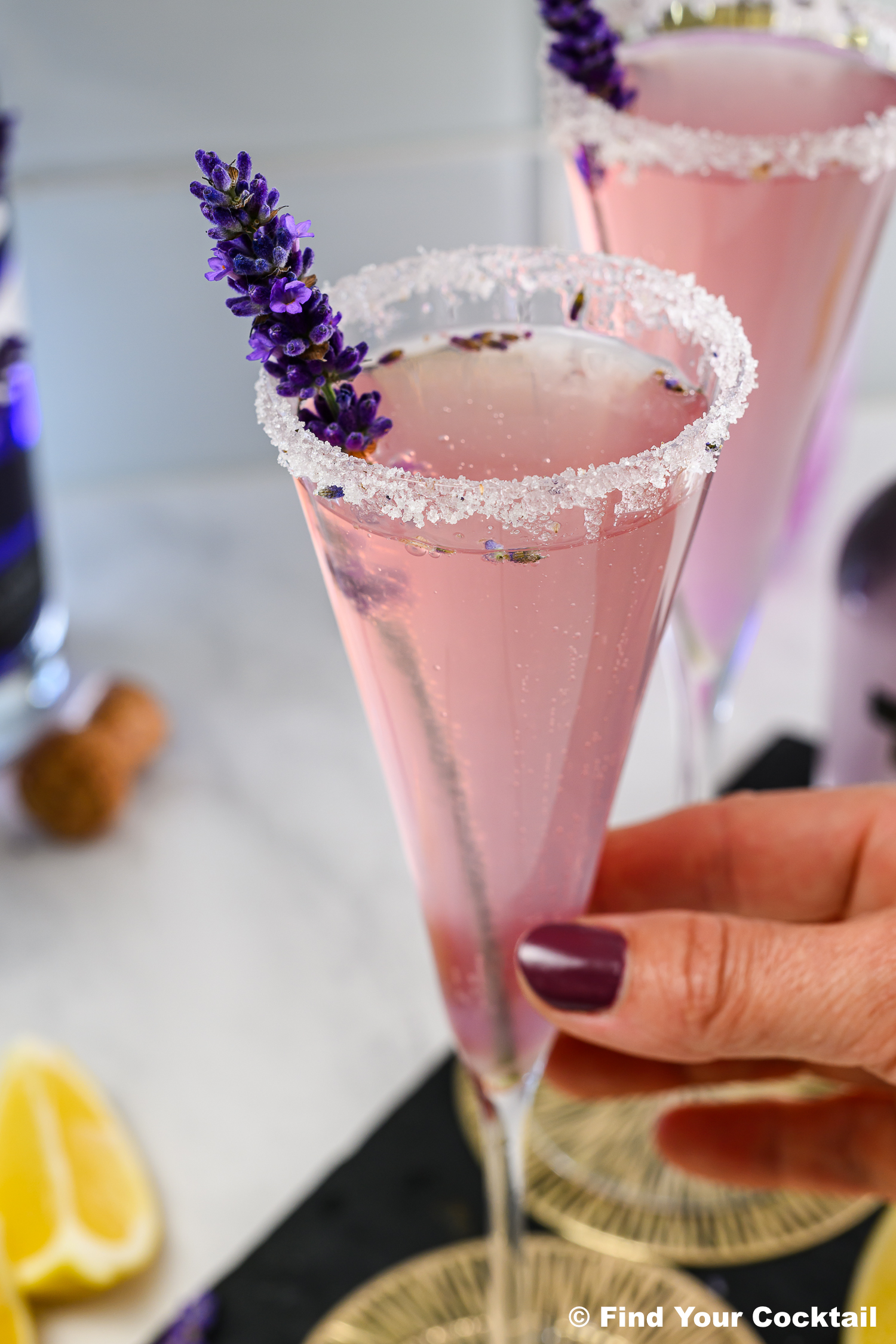 A hand holding a champagne flute with a pink cocktail, sugar-rimmed glass, and a sprig of lavender garnish; lemon wedges and a cork are in the background.