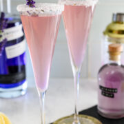 Two pink cocktails in champagne flutes with sugared rims and lavender sprigs, set on a counter with bottles, a shaker, and lemon wedges in the background.