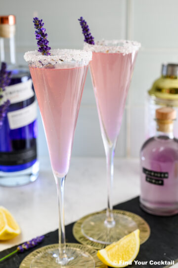 Two pink cocktails in champagne flutes with sugared rims and lavender sprigs, set on a counter with bottles, a shaker, and lemon wedges in the background.