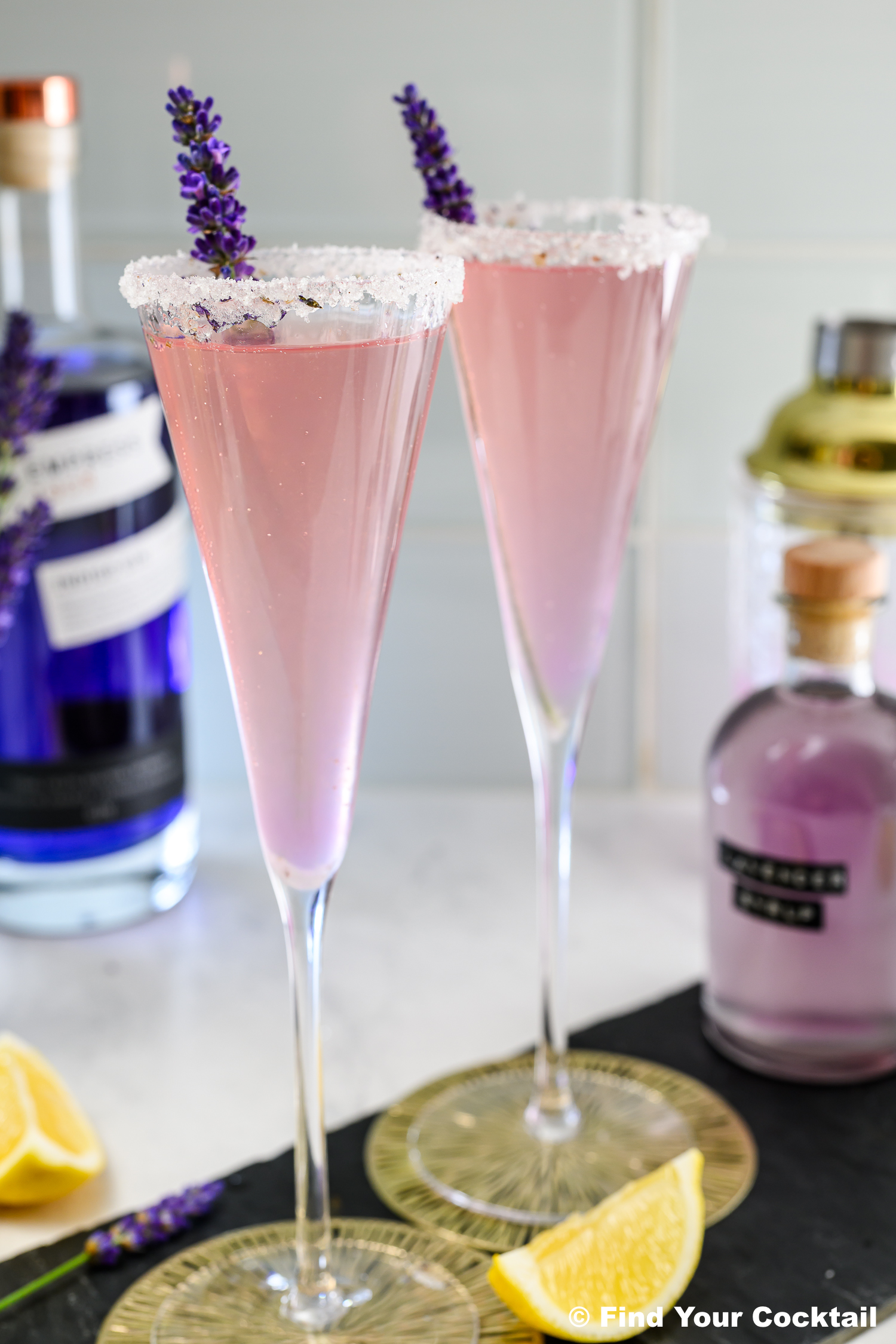 Two pink cocktails in champagne flutes with sugared rims and lavender sprigs, set on a counter with bottles, a shaker, and lemon wedges in the background.
