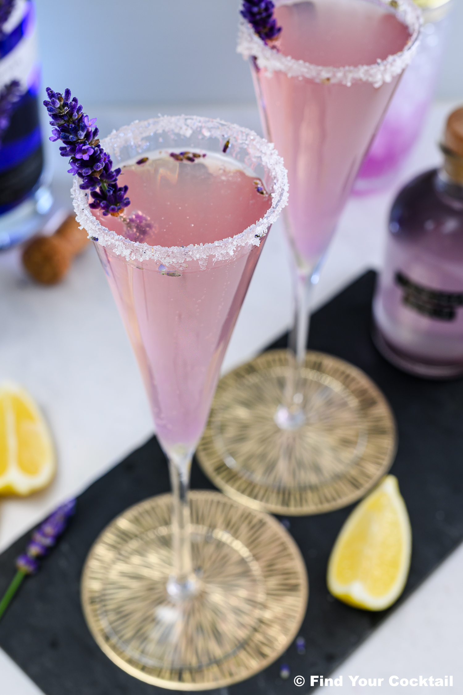 Two pink cocktails in champagne flutes with sugared rims, garnished with lavender sprigs, placed on gold-patterned coasters next to lemon wedges.