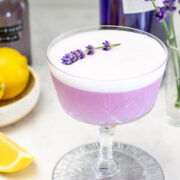 A purple cocktail with a frothy top, garnished with a sprig of lavender, is served in a coupe glass. Lemons and bottles are visible in the background.