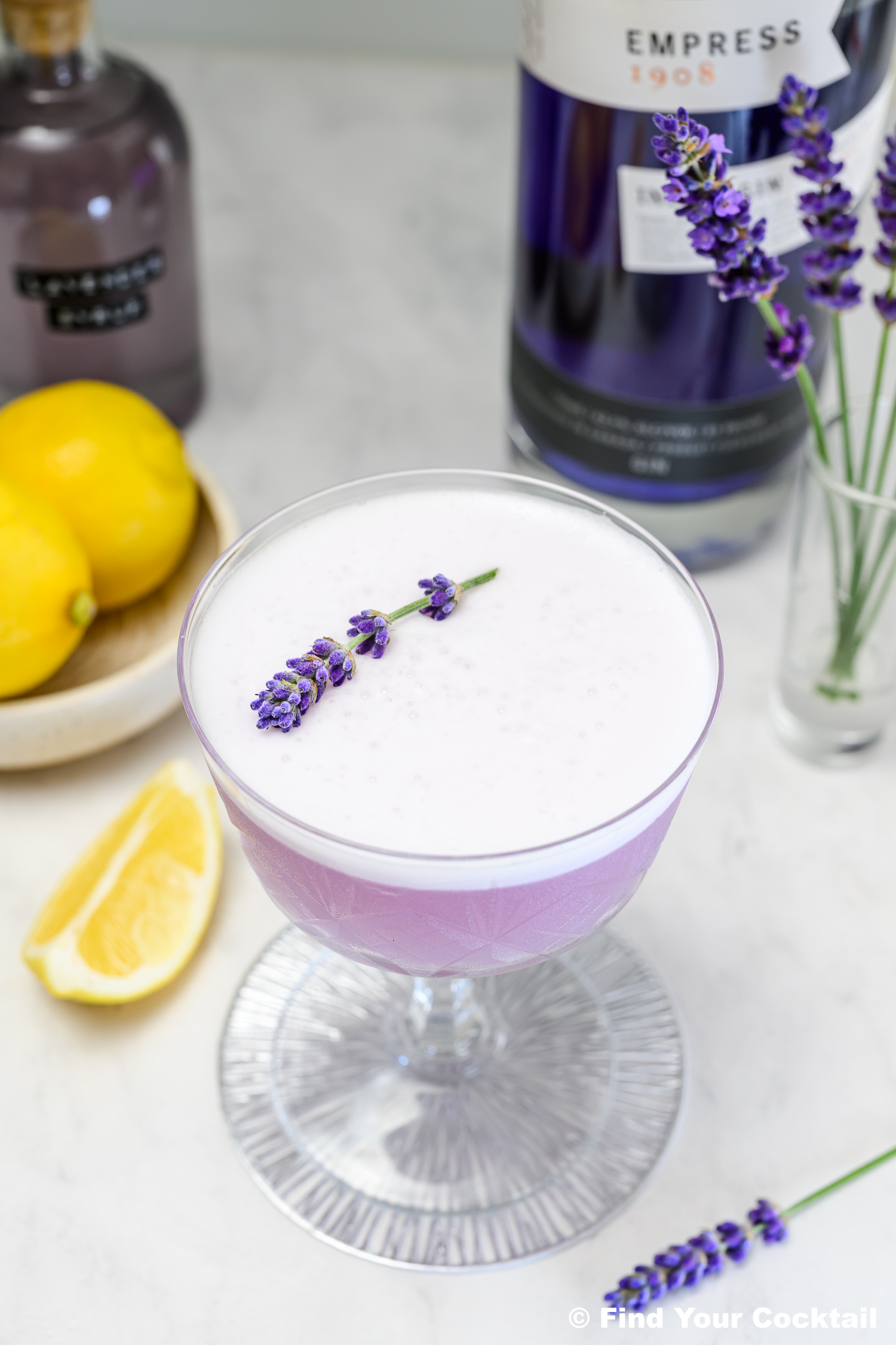A purple lavender cocktail in a coupe glass garnished with a lavender sprig, with lemons, gin bottles, and a glass vase with lavender in the background.