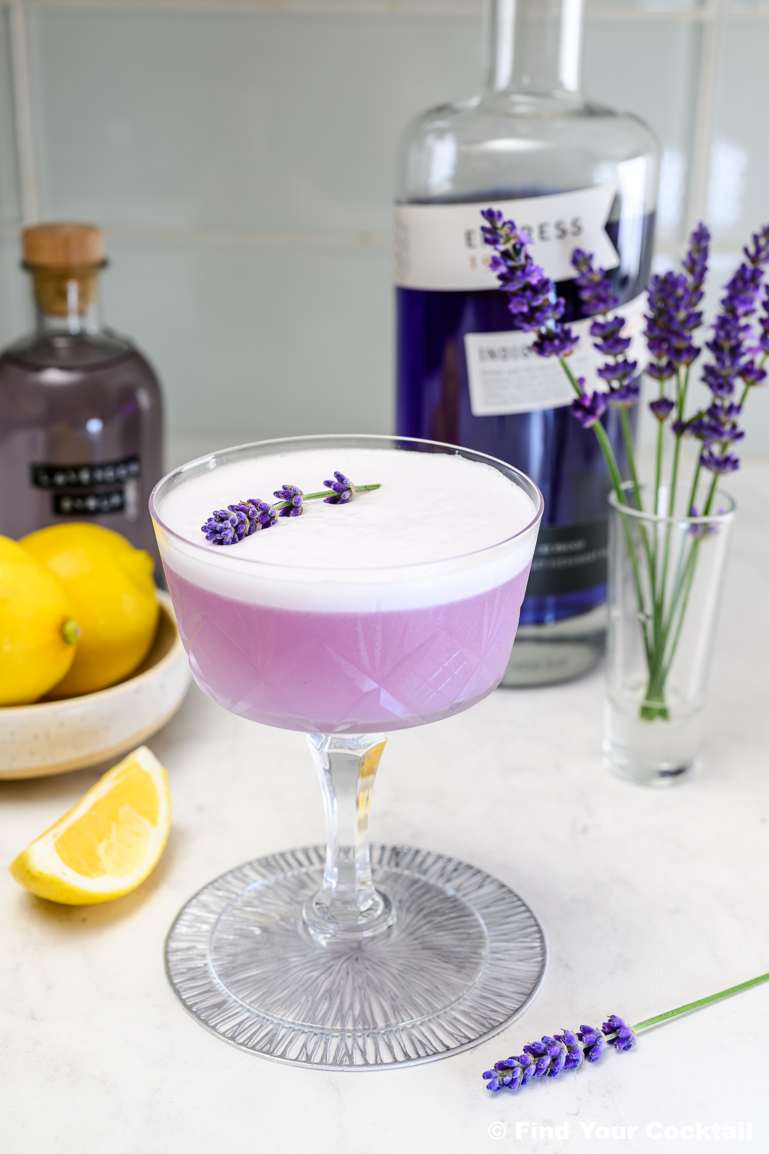 A purple cocktail with a frothy top is garnished with lavender in a coupe glass, surrounded by lemons, a bottle of gin, lavender sprigs, and a glass vase on a countertop.
