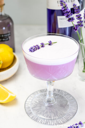 A purple cocktail with a frothy top, garnished with a sprig of lavender, is served in a coupe glass. Lemons and bottles are visible in the background.