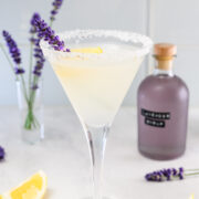 A martini glass with a sugar rim, filled with a pale cocktail, garnished with a lemon slice and lavender sprig; a purple bottle and lemon wedges are nearby.