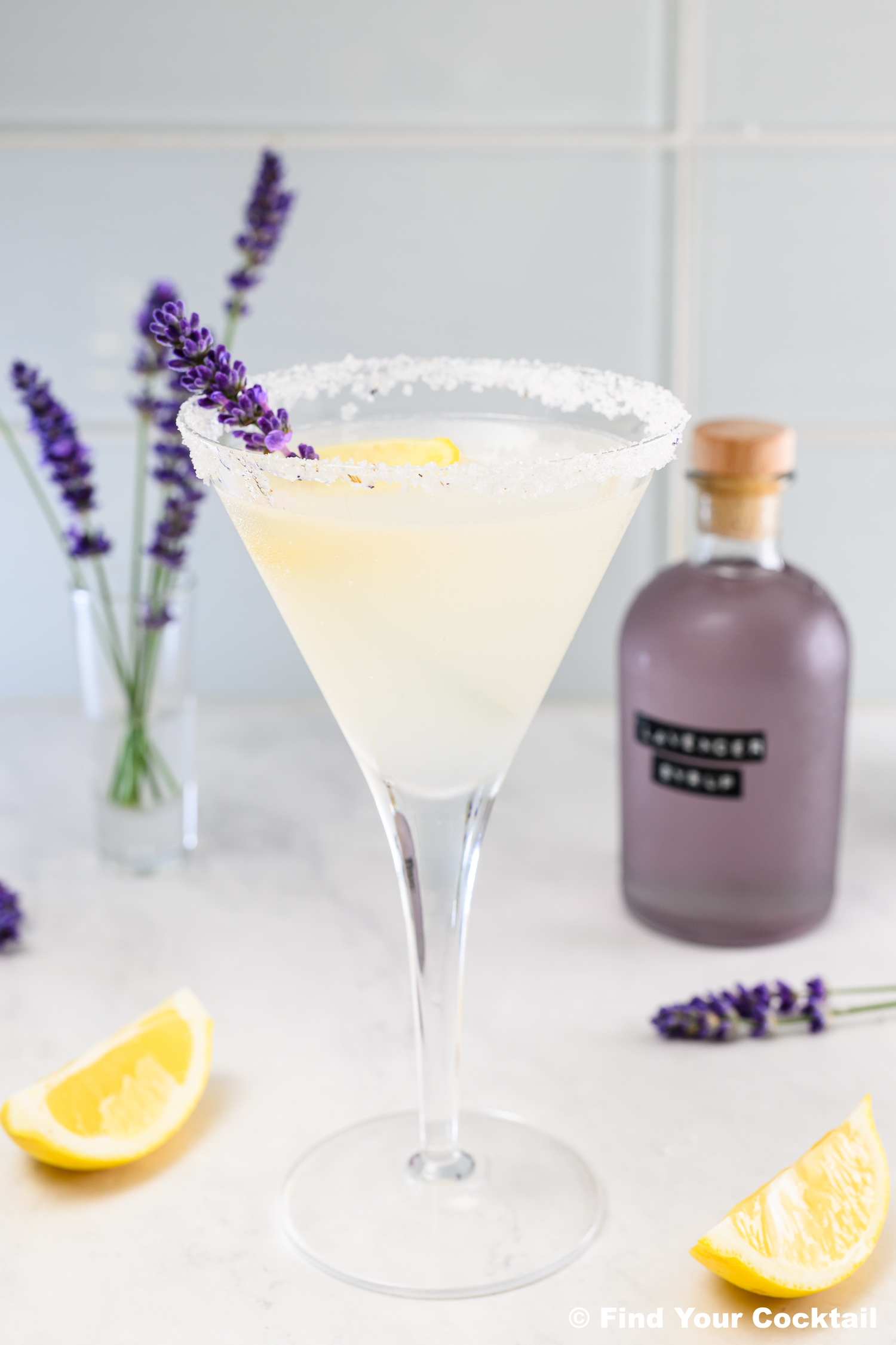 A martini glass with a sugar rim, filled with a pale cocktail, garnished with a lemon slice and lavender sprig; a purple bottle and lemon wedges are nearby.