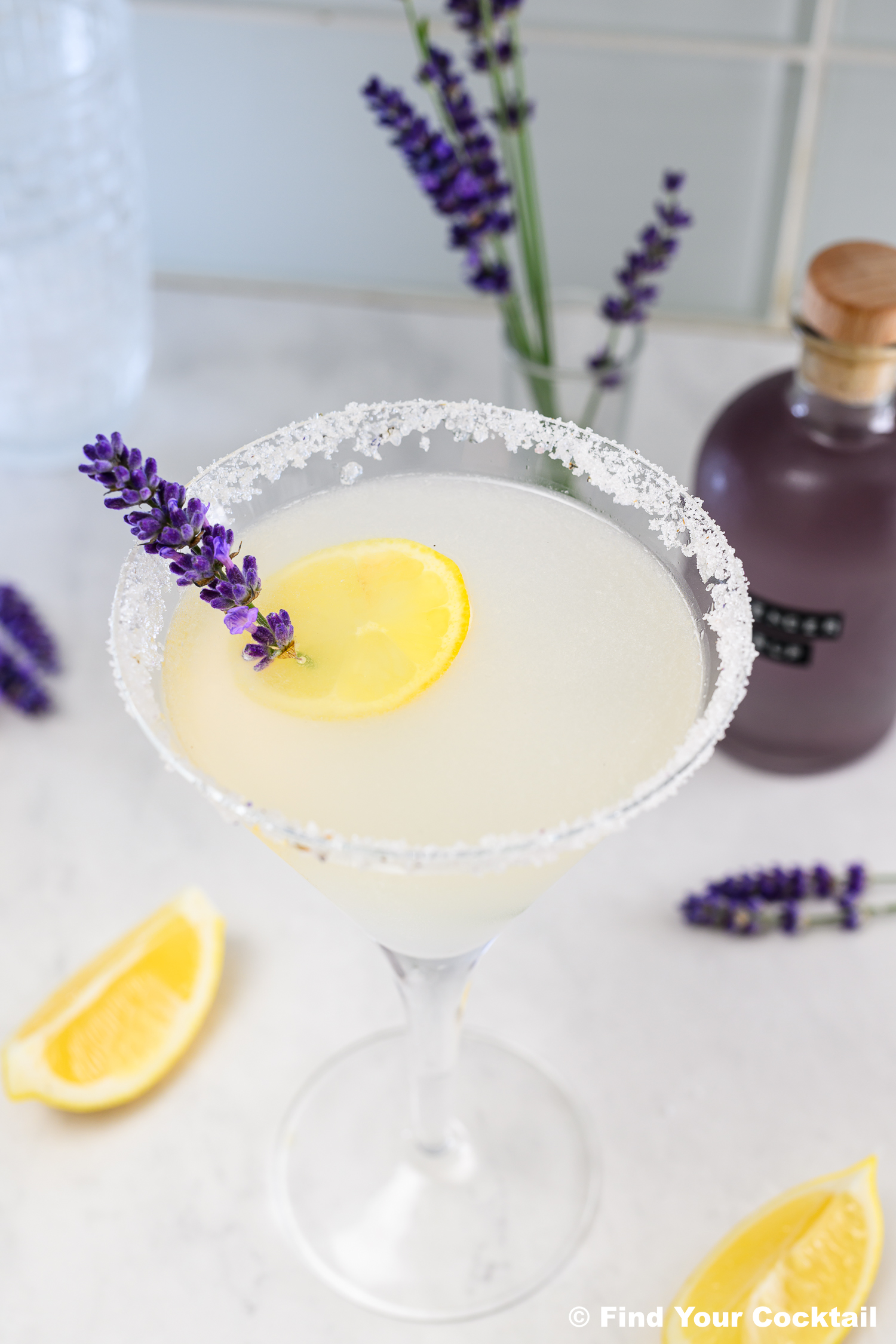 A cocktail in a martini glass with a salted rim, garnished with a lemon slice and a sprig of lavender, with lavender flowers and lemon wedges nearby.