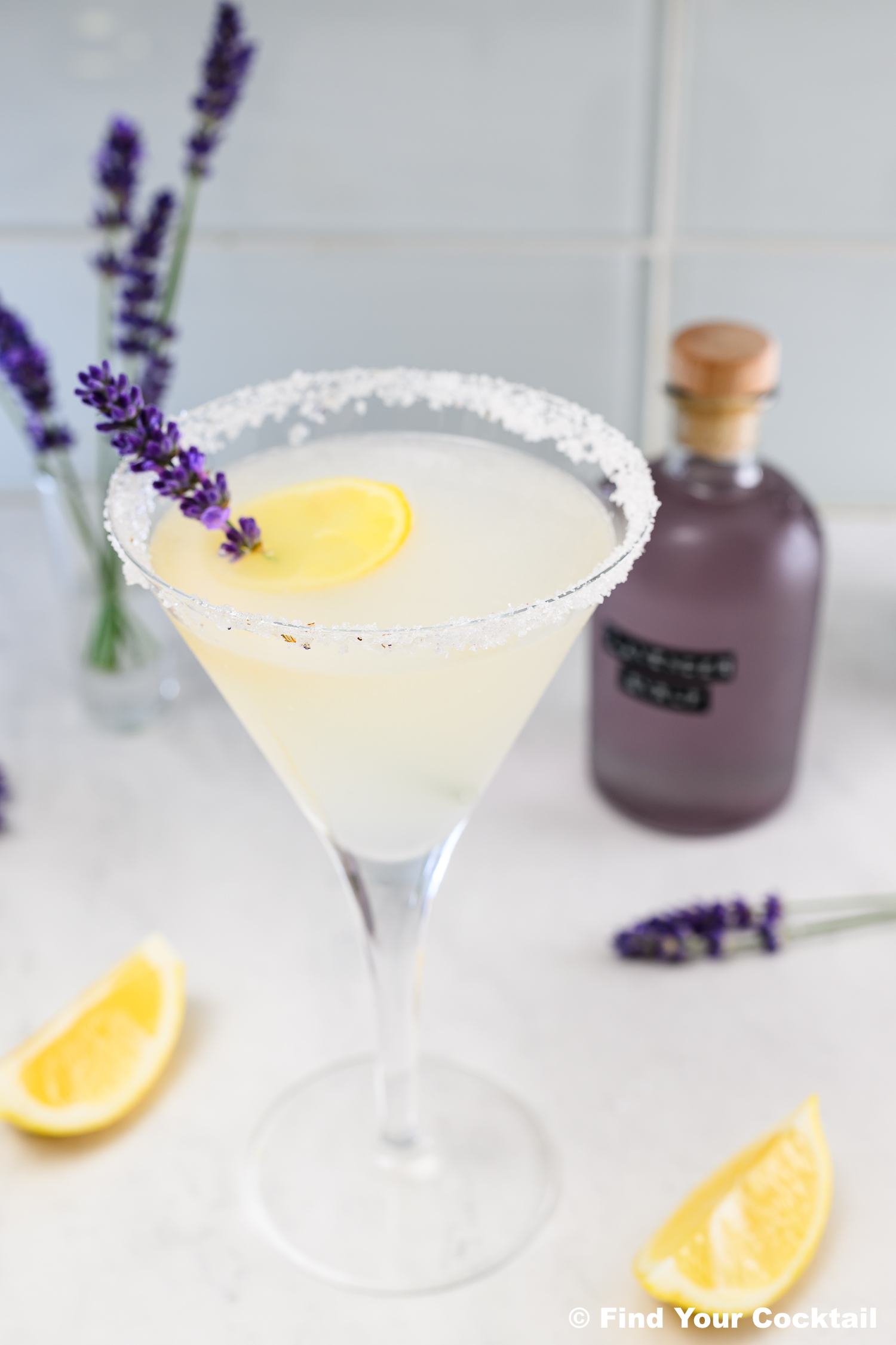 A martini glass with a salted rim, filled with a pale yellow cocktail, garnished with a lemon slice and lavender sprig. Lavender flowers and a bottle are in the background.