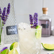 A glass of iced cocktail garnished with lavender sprigs and a lime wedge sits on a table with cut limes, lavender flowers, and bottles of tequila and syrup in the background.