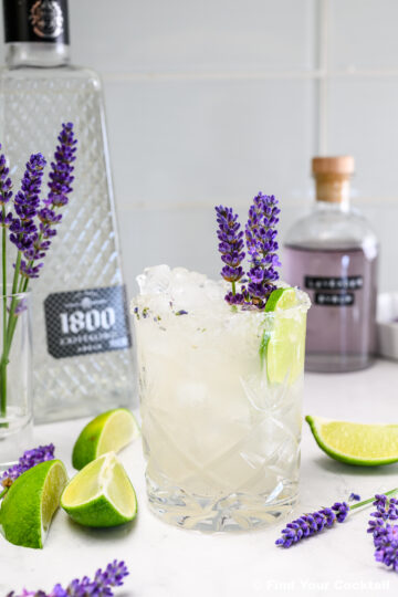 A glass of iced cocktail garnished with lavender sprigs and a lime wedge sits on a table with cut limes, lavender flowers, and bottles of tequila and syrup in the background.