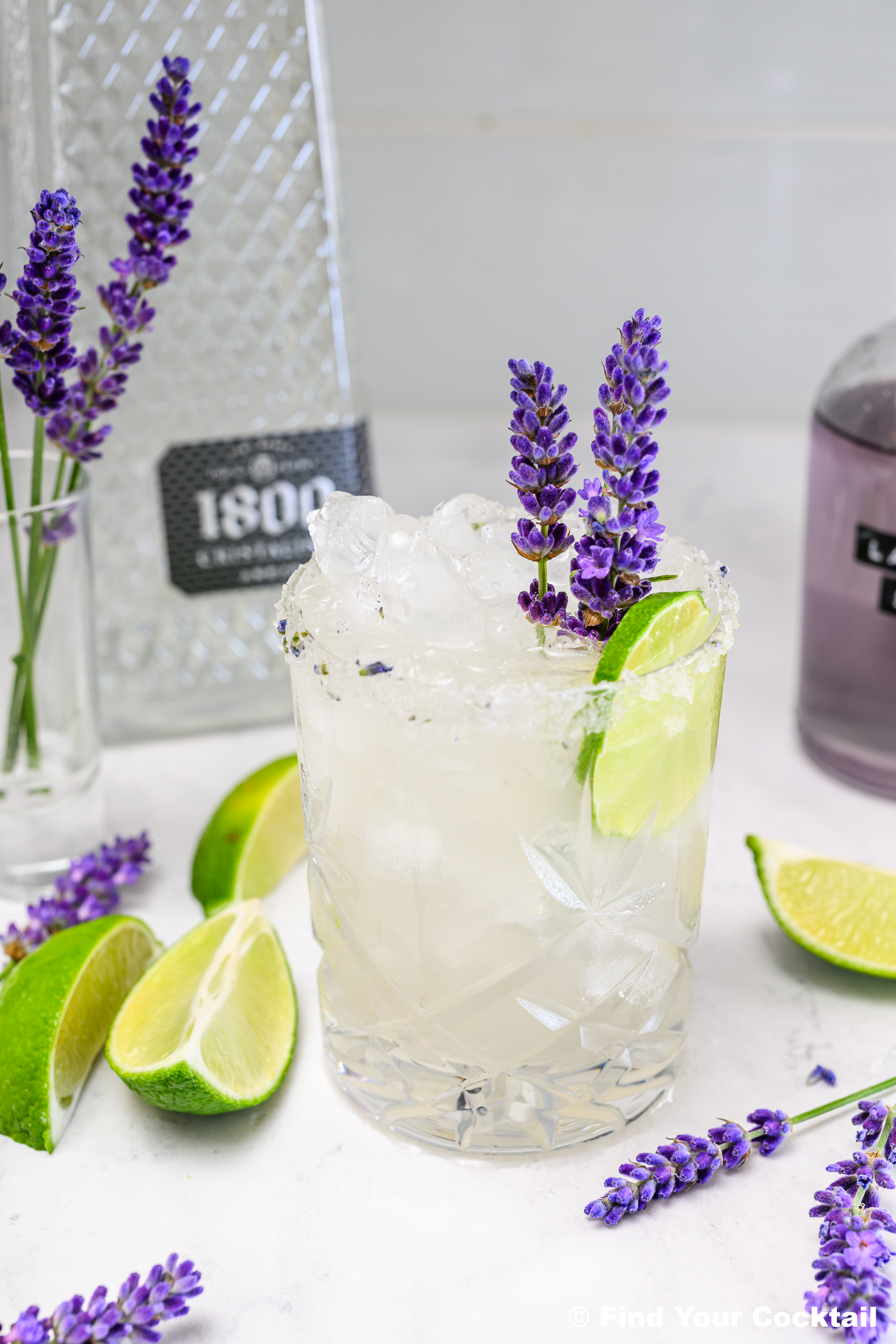 A glass of iced cocktail garnished with a lime wedge and sprigs of lavender, with cut limes and lavender flowers on the table beside bottles in the background.