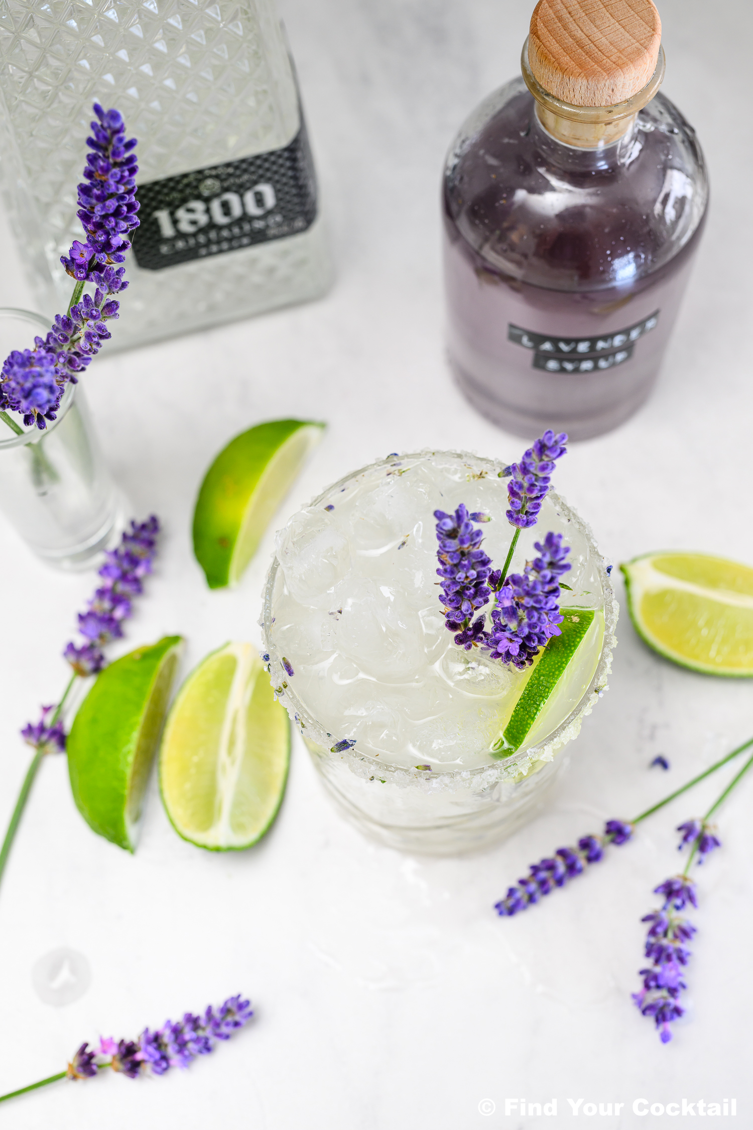 A glass of iced cocktail garnished with lavender and lime, surrounded by lime wedges, lavender sprigs, a bottle of tequila, and a bottle labeled lavender syrup on a white surface.
