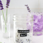 A glass jar labeled Lavender Salt sits on a white counter, surrounded by fresh lavender sprigs and a glass of lavender flowers in the background.
